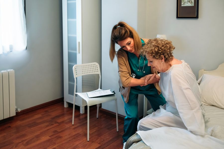 image presents nurse helping elder woman to stand in the bed